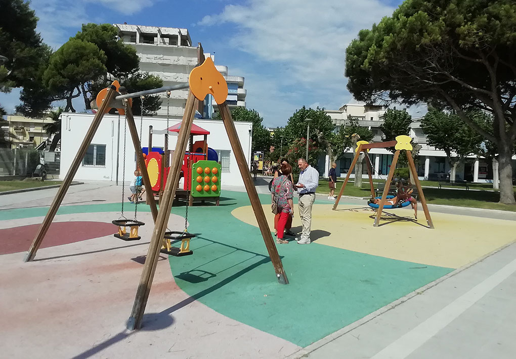 un'area gioco con altalene, di cui una inclusiva, e scivoli. AI lati dell'area si vedono alberi e sullo sfondo edifici