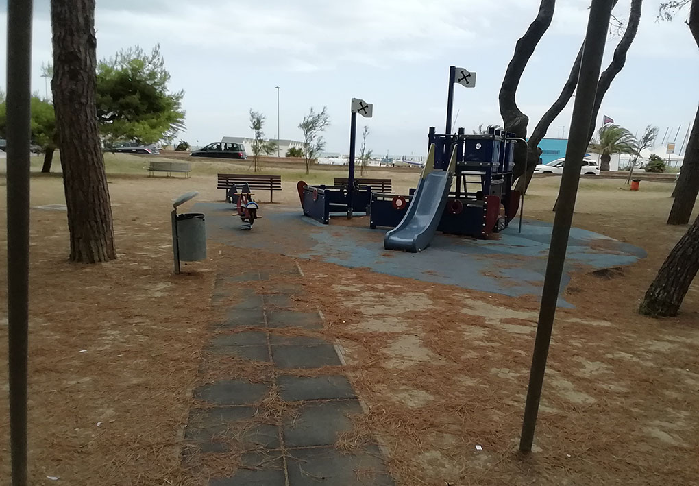 Giochi tra gli alberi della pineta. Un vialetto in gomma antitrauma arriva alla zona giochi, sullo sfondo il lungomare, rialzato rispetto al parco.