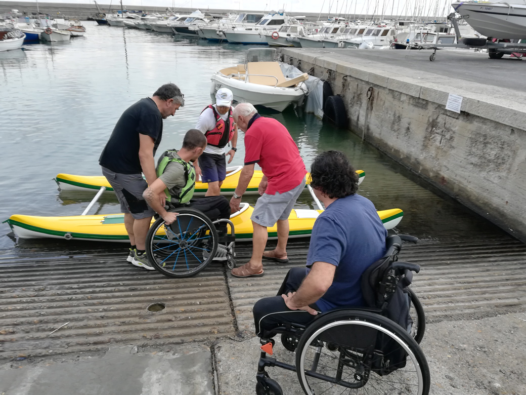 Una persona in carrozzina viene aiutata a salire su una canoa doppia, mentre un'altra persona in carrozzina li guarda