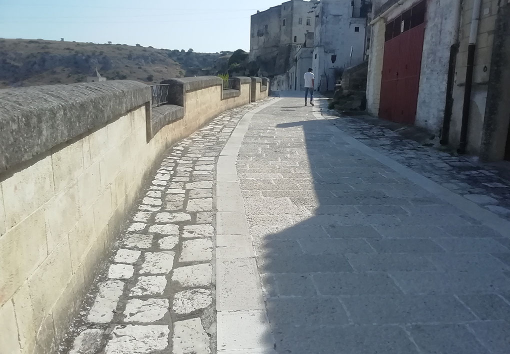 un'ampia strada lastricata con edifici in pietra da un lato e un muraglione che, si intuisce, si affaccia sulla vallata dall'altro.