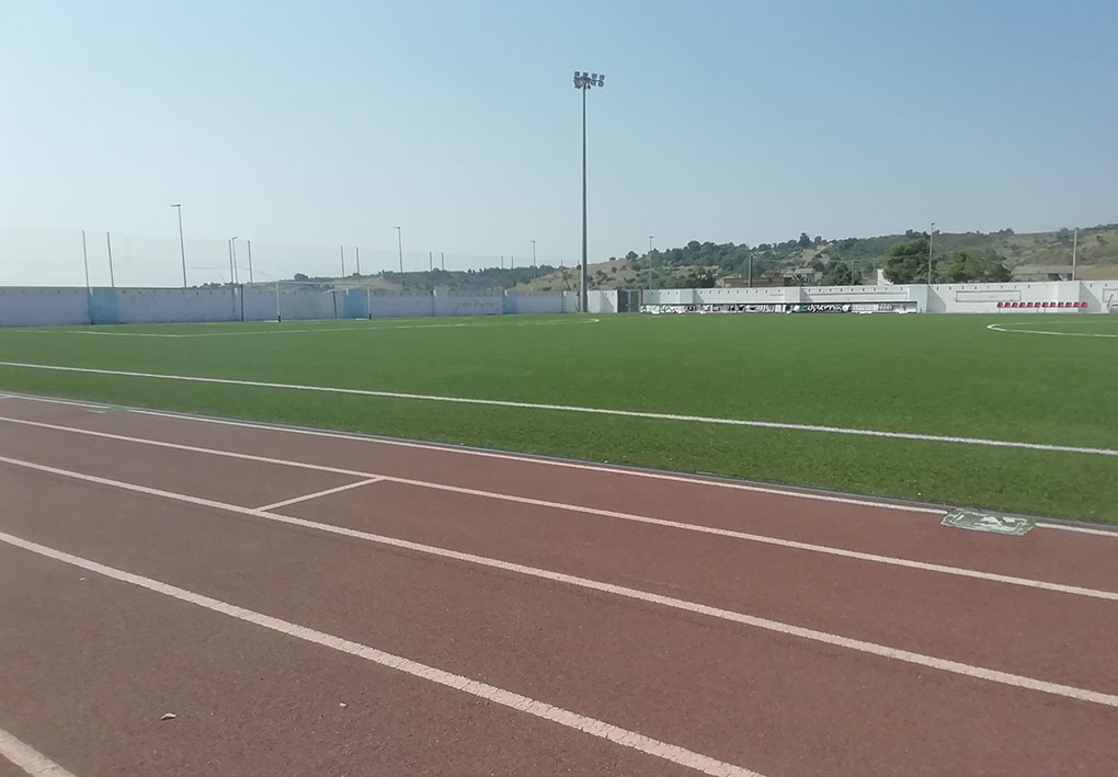 Pista da corsa in terra intorno a un campo da gioco in erba. Sullo sfondo le panchine per gli atleti