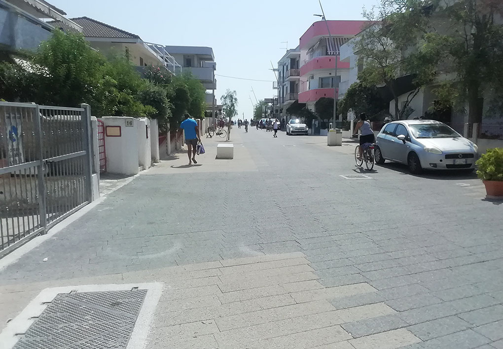 Un'ampia strada piastrellata tra basse palazzine e alberi ai lati. Si notano poche macchine parcheggiate e alcune persone in bicicletta