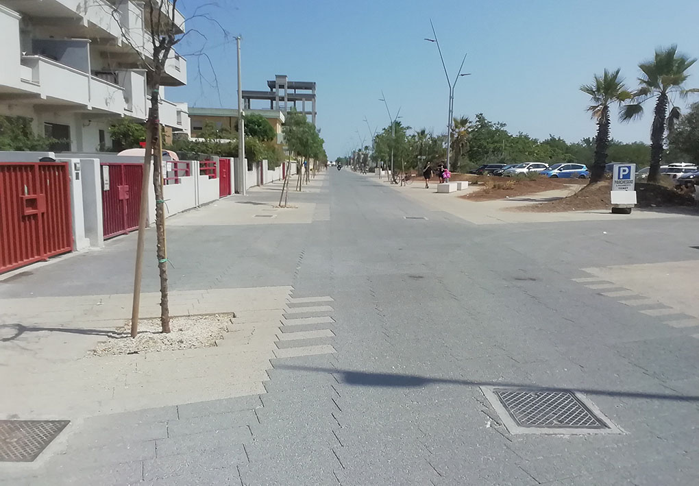 un'ampia strada, da un lato basse palazzine e dall'altro palme e un parcheggio.