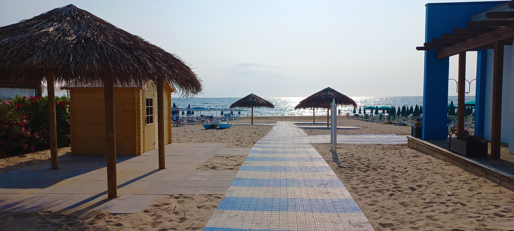 Una passerella si allunga sulla spiaggia sabbiosa, tra ombrelloni di paglia