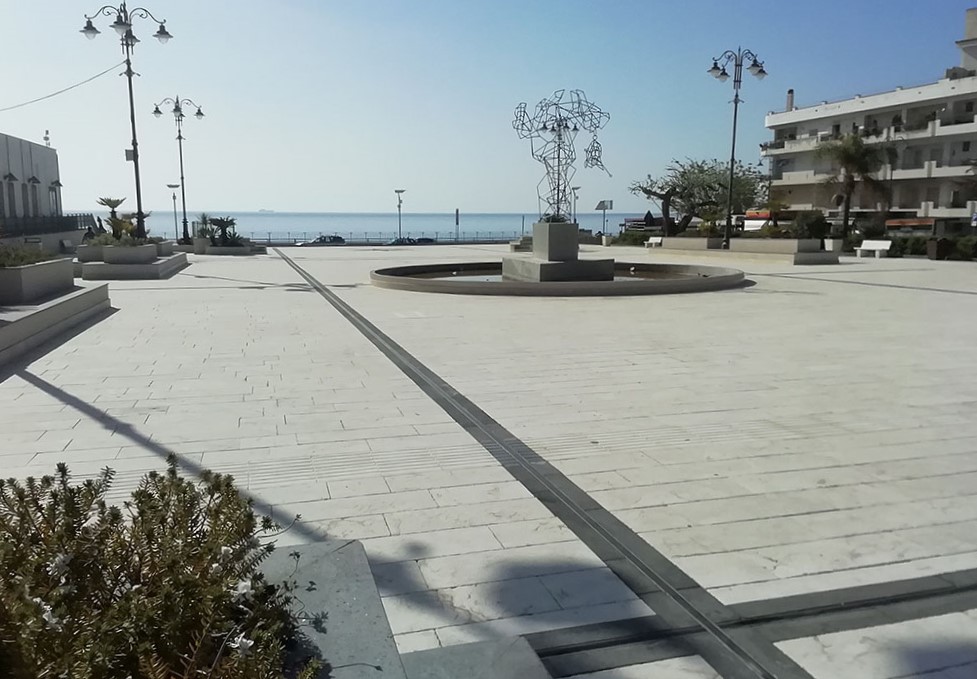 Un'ampia piazza pianeggiante, ai suoi lati ci sono bassi palazzi e sullo sfondo il mare. Al centro della piazza sorge un monumento e sul perimetro sorgono aiuole.