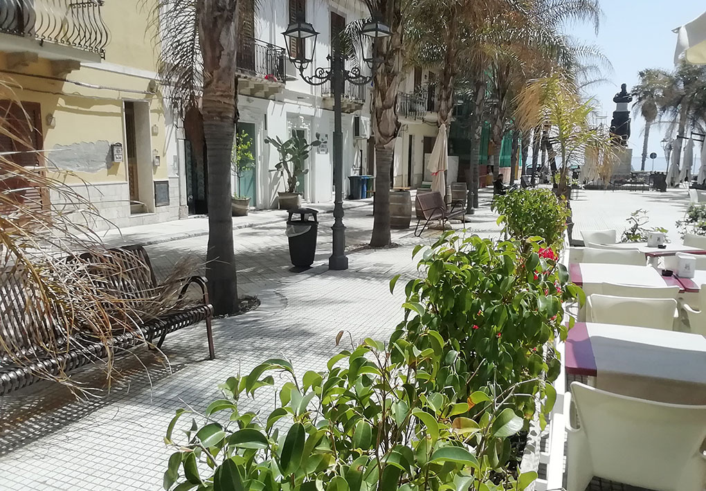 una piazza pianeggiante con palme, panchine e aiuole. Sullo sfondo, oltre un monumento, il mare