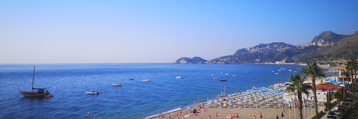 panorama sulla costa. Sul fondo un promontorio si allunga sul mare. In primo piano ombrelloni di stabilimenti punteggiano la spiaggia di sabbia 