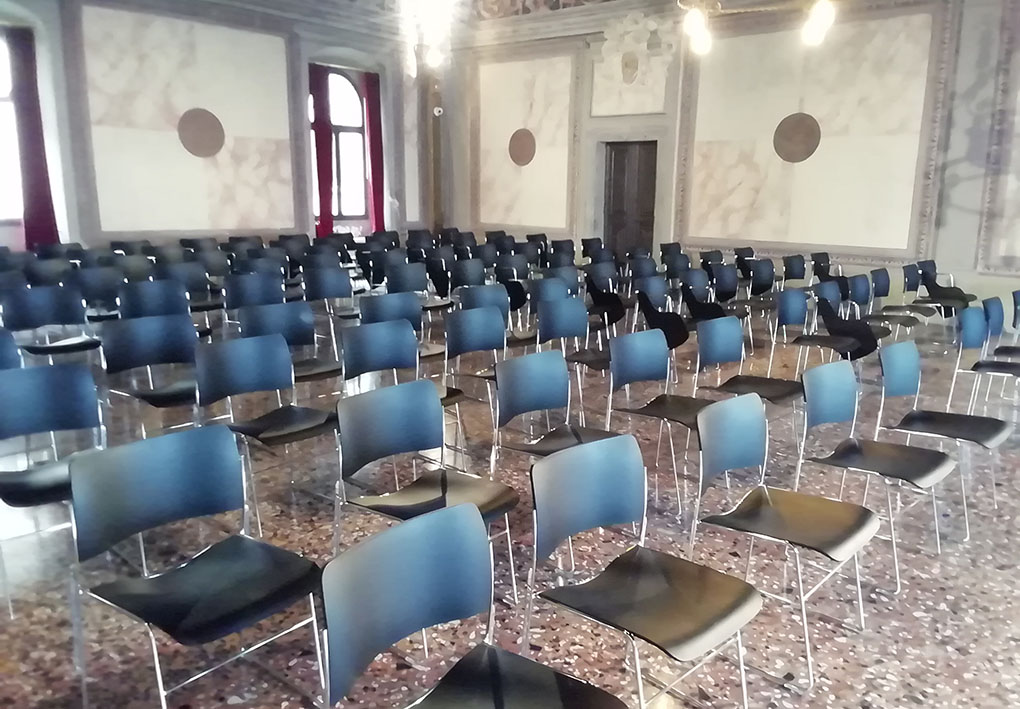 in una sala di un palazzo storico, numerose file di sedie compongono una platea