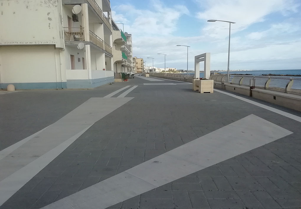 ampia strada piastrellata in riva al mare. dal lato opposto alla spiaggia, palazzi moderni si affacciano direttamente sulla via. Lungo la strada, una scultura moderna arreda lo spazio.