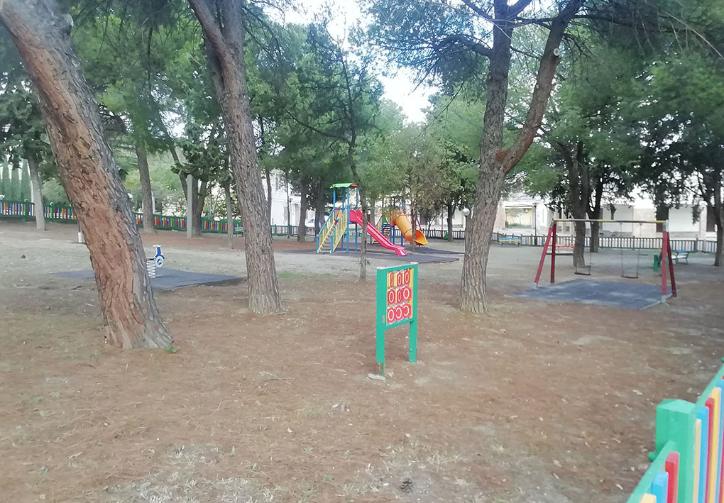 Area giochi per bambini tra gli alberi. In un angolo si nota la recinzione in legno colorato che delimita l'area.