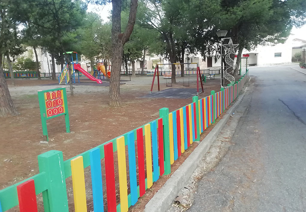 Strada asfaltata a fianco del parco giochi, racchiuso da uno steccato colorato. Gli alberi del parco fanno ombra anche sulla strada carrozzabile