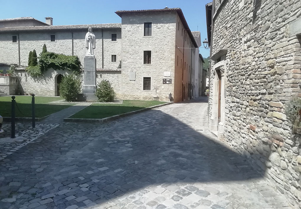 Una via lastricata tra palazzi storici in pietra. Un'area verde si apre sul lato della strada, al suo centro è eretta una statua.