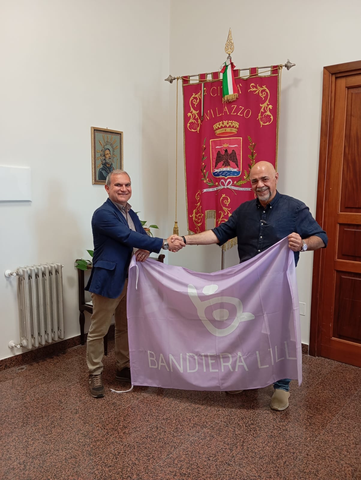 Il presidente Roberto Bazzano posa con il sindaco di Milazzo davanti al gonfalone della città. Con una mano tengono la bandiera, con l'altra si dannò una stretta di mano.