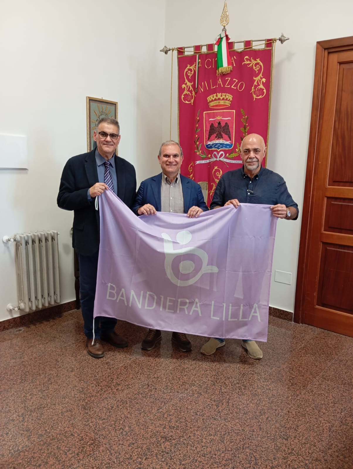 Il presidente di Bandiera Lilla, il sindaco e l'assessore di Milazzo posano di fronte al gonfalone della Città, mostrando la Bandiera Lilla