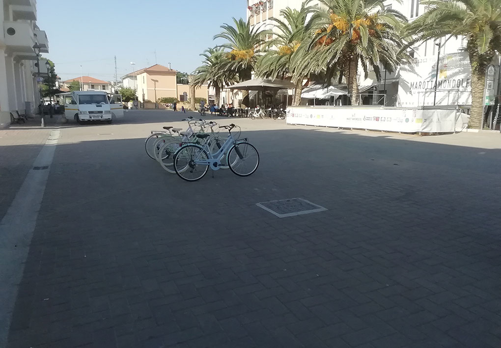 piazza pedonale con palme e alcune biciclette parcheggiate. A un lato della piazza è montato un palco.