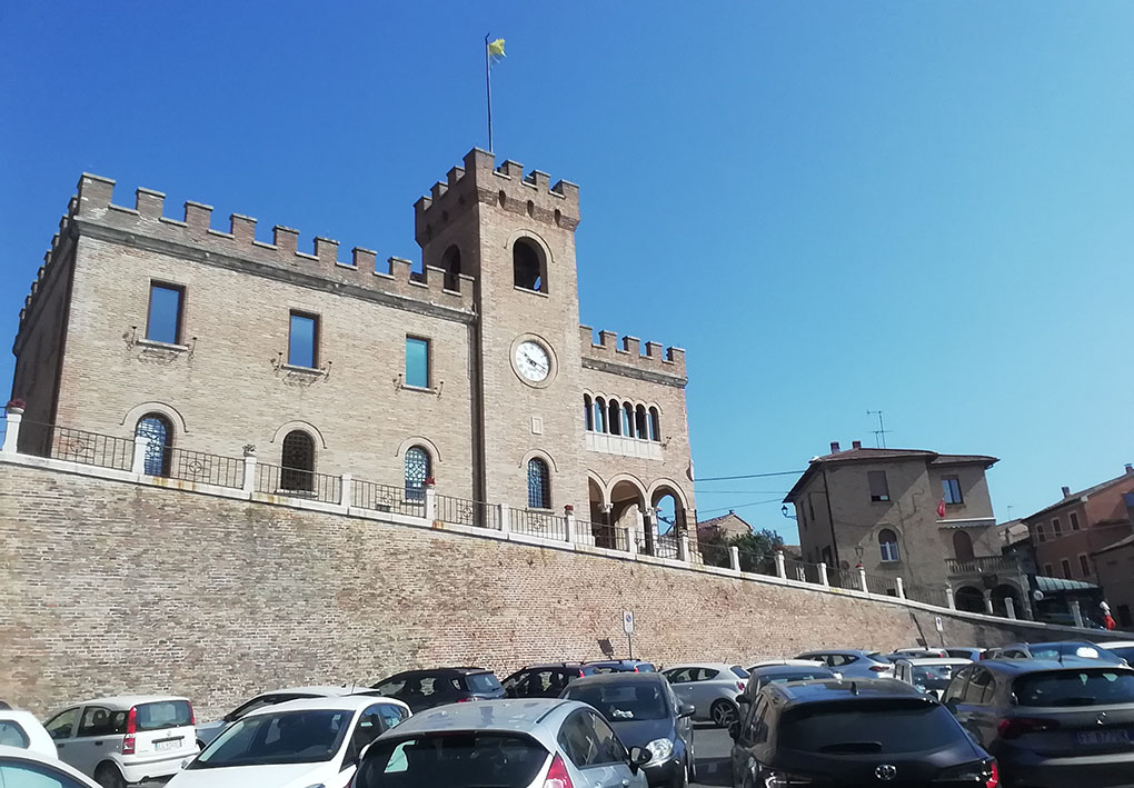 Lo storico palazzo comunale, in mattoni, visto dall'ampio parcheggio sottostante. Una strada in salita mediamente ripida sale verso il palazzo.