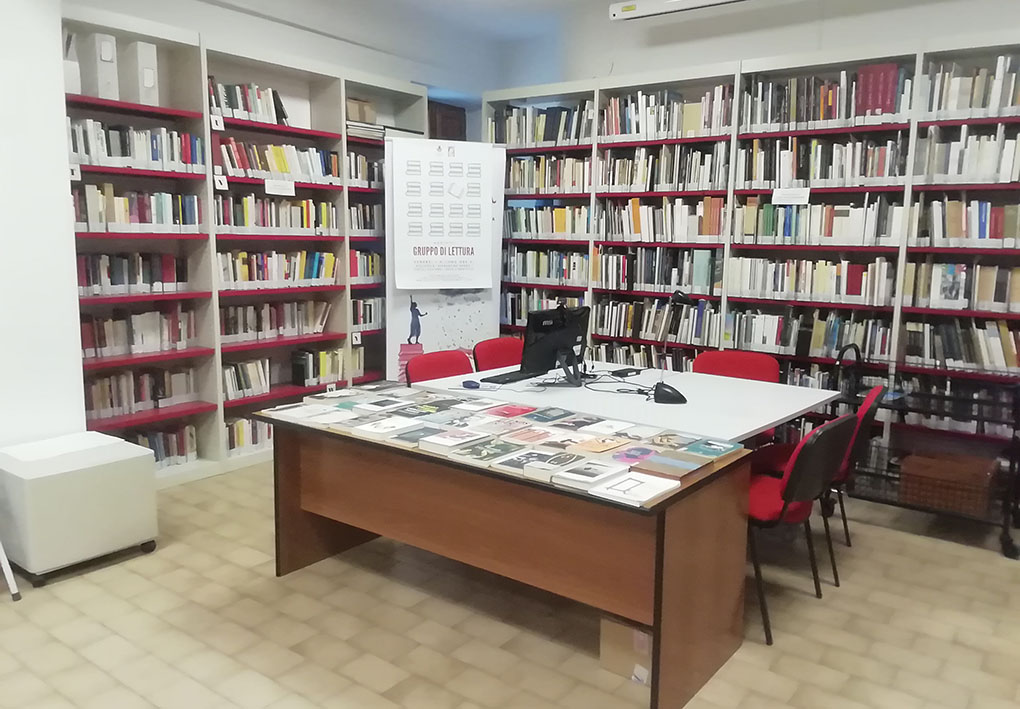 Sala con postazione internet. In una sala con fitti scaffali alle pareti, un tavolo con 2 sedie per lato ospita un computer. A fianco al tavolo, una cattedra espone numerosi libri.