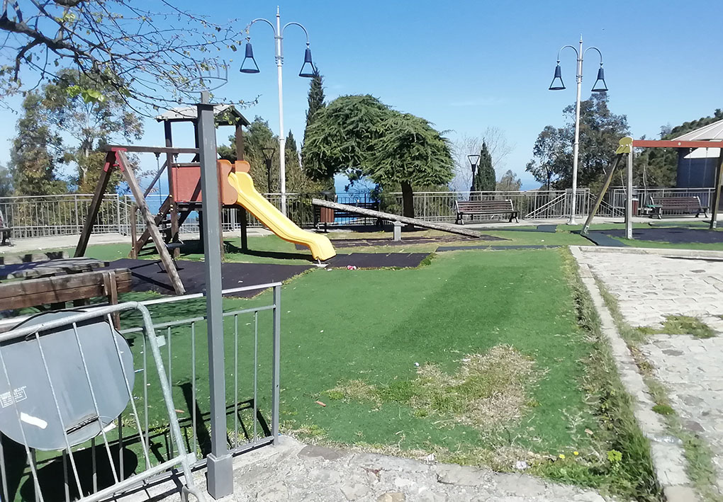 Un parco giochi in un'ampia area. Tra el varie attrezzature, uno scivolo e delle altalene. Sul lato un vialetto in pietra delimita l'area gioco