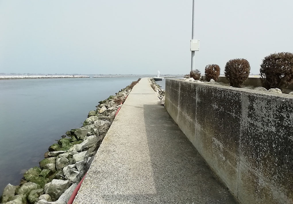 Una strada di cemento si inoltra nella laguna. Ai lati scogli la riparano dalle onde. All'inizio dell'inquadratura, una fioriera di cemento fa da parete laterale.