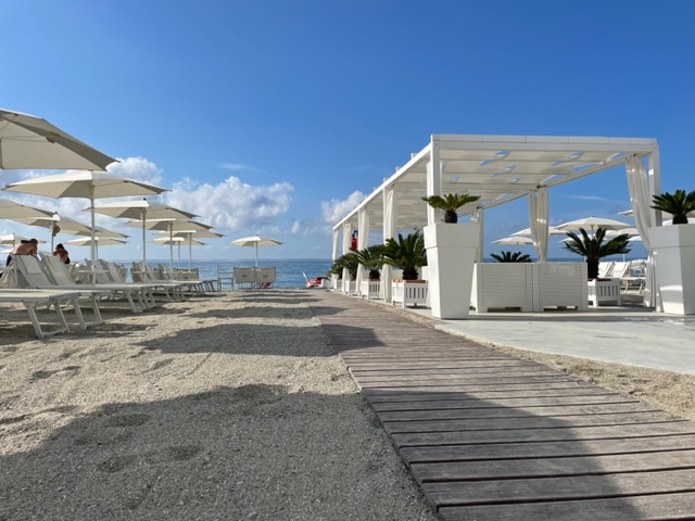 Una spiaggia di sabbia con ombrelloni chiari e una tettoia. Una passerella di legno si allunga verso il mare